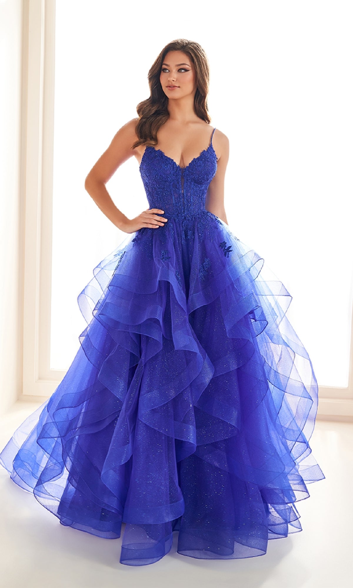 A woman stands in a bright room wearing the Ellie Wilde EW36203 Glitter Tulle Prom Ball Gown, featuring deep blue glitter tulle, thin straps, and a fitted embroidered corset bodice.
